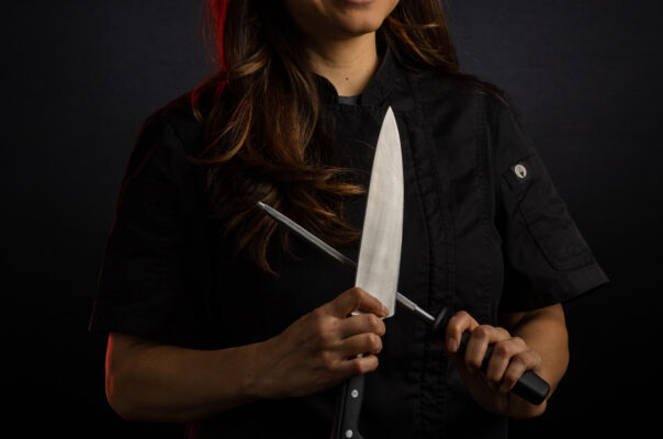 Female chef in black uniform holding knife and sharpener in front of her with high contrast lighting.