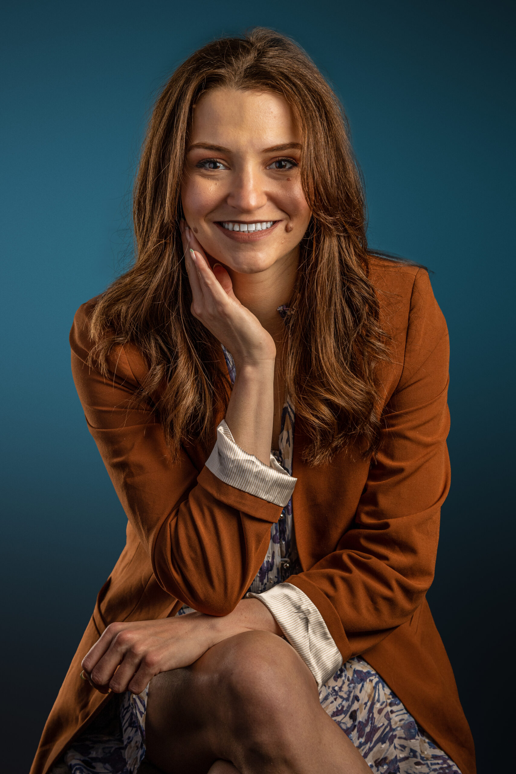 Brunette woman professional in rust colored jacket sitting cross legged with elbow on knee and hand on side of face in front of teal background.