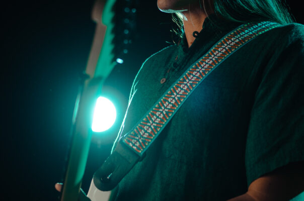 Waist up shot of male guitar player with long hair playing a white Fender Stratocaster while getting bathed in turquoise light