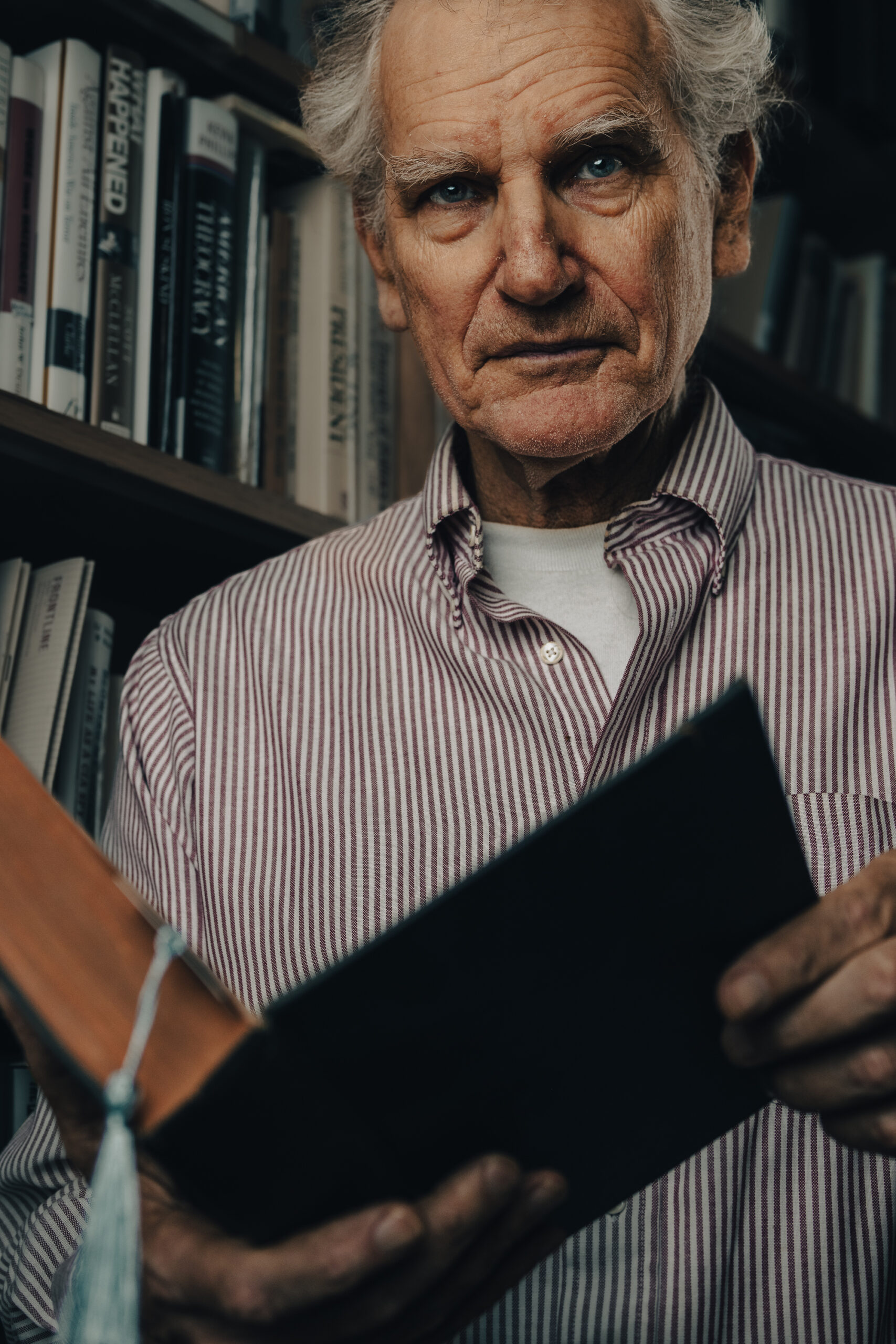 Close up shot of older man in light colored collared shirt standing in front of a library shelf looks up from his book.