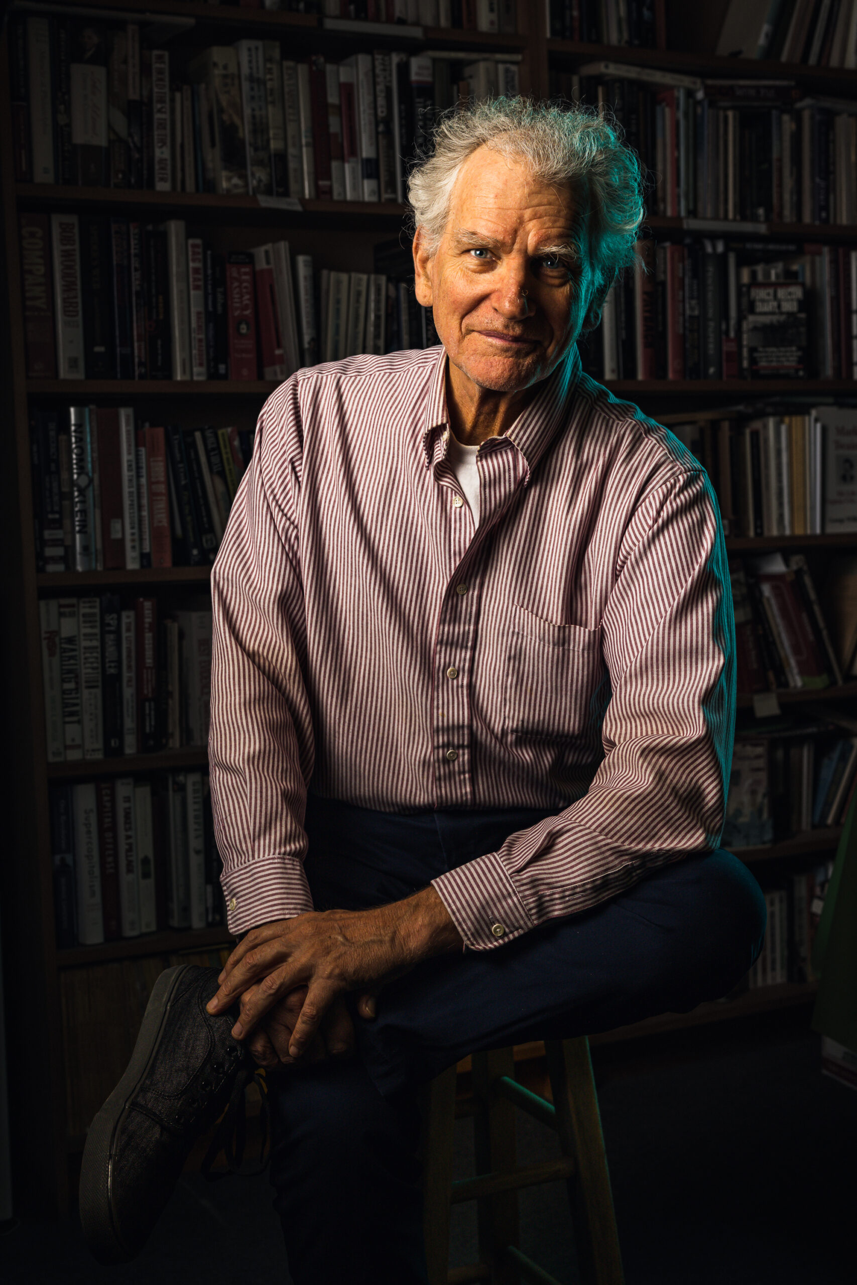 Older man in light colored collared shirt sits on a stool while leaning towards the camera while being illuminated on the left side by a warm light, and a cool turquoise light from the opposite side.