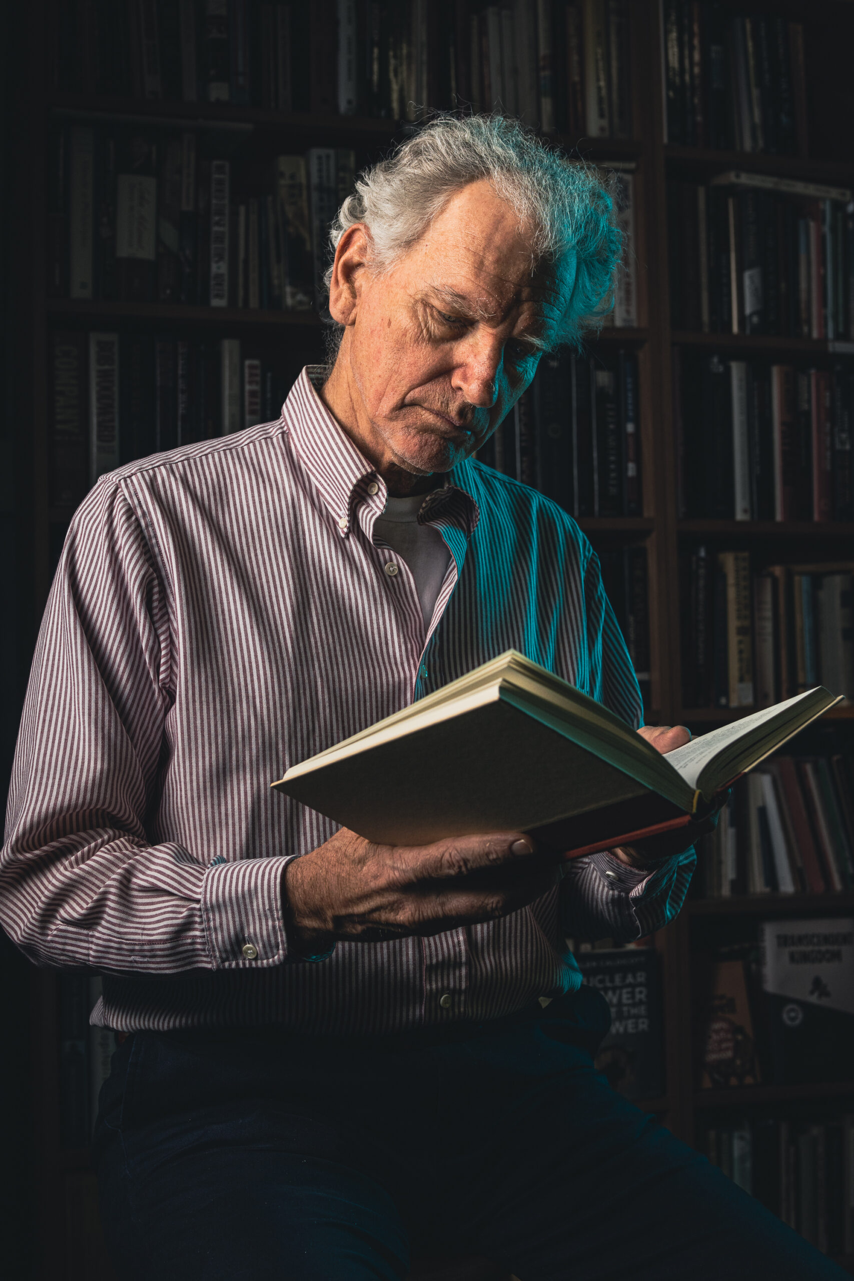 Older man in light colored collared shirt reads a book in front of darkened library shelf while he is bathed in warm light on one side and turquoise light from the opposite side.
