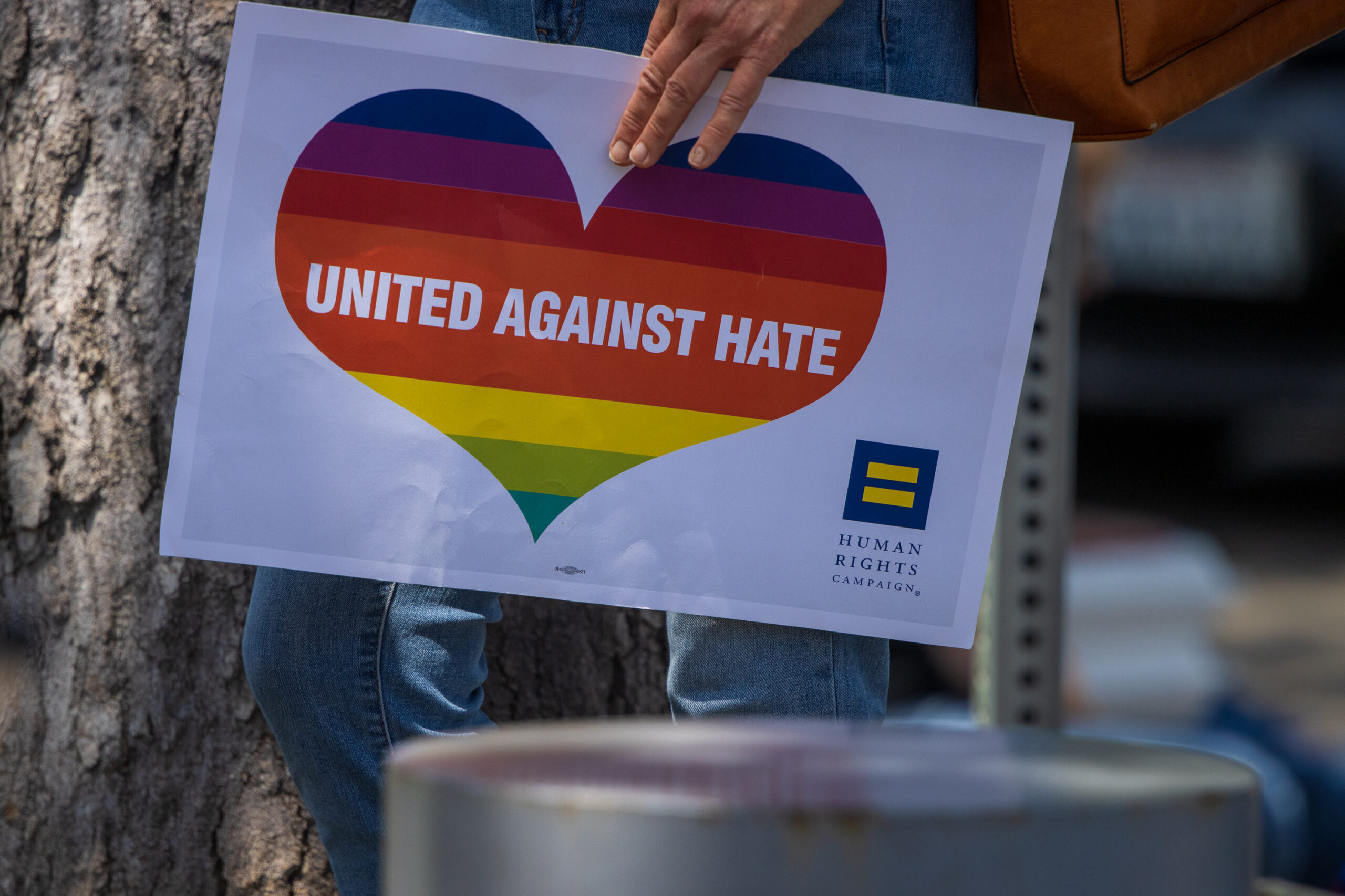 Person wearing jeans leaning against a tree in the shade holds a light grey sign with a rainbow heart in the center which reads, "United Against Hate."