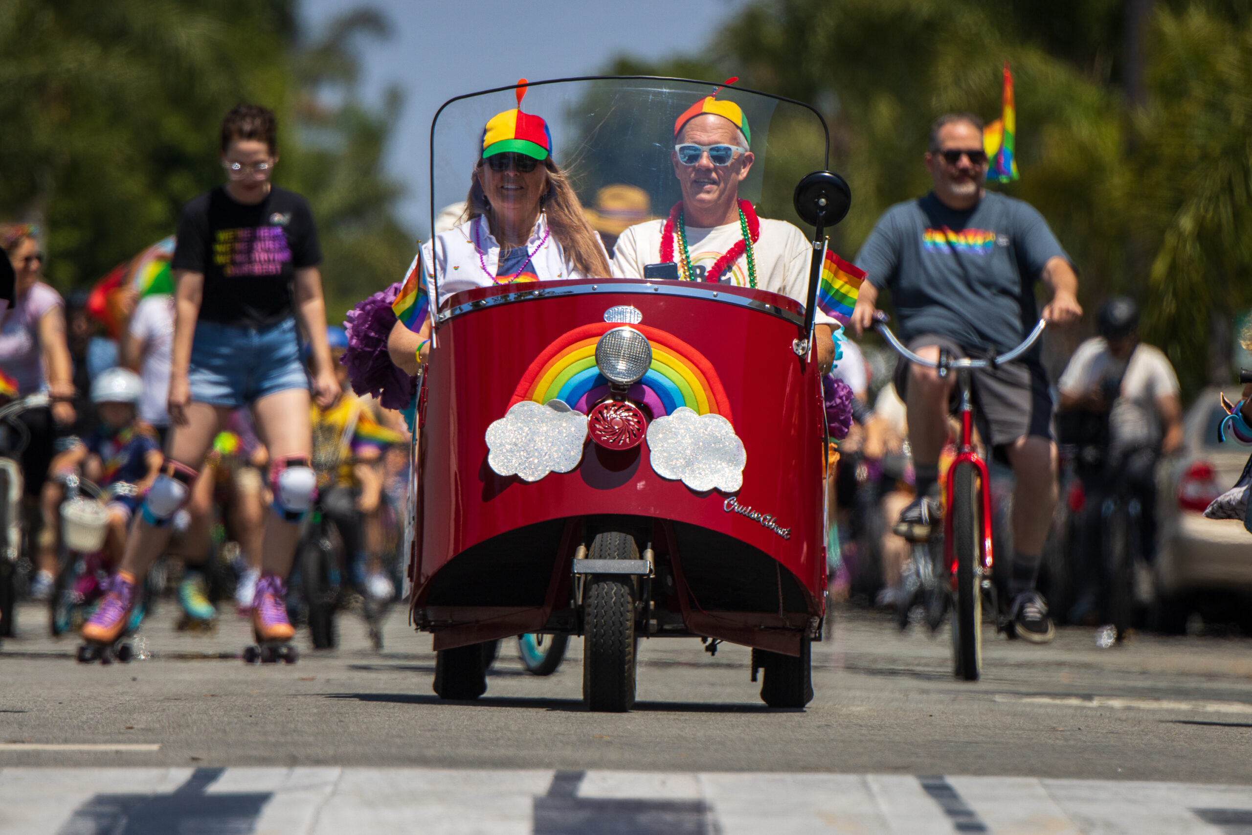 Deep candy red convertible tiny car with rainbow decoration leads the Fullerton Pride Fest 2023 bicycle parade into the Fullerton Museum plaza.