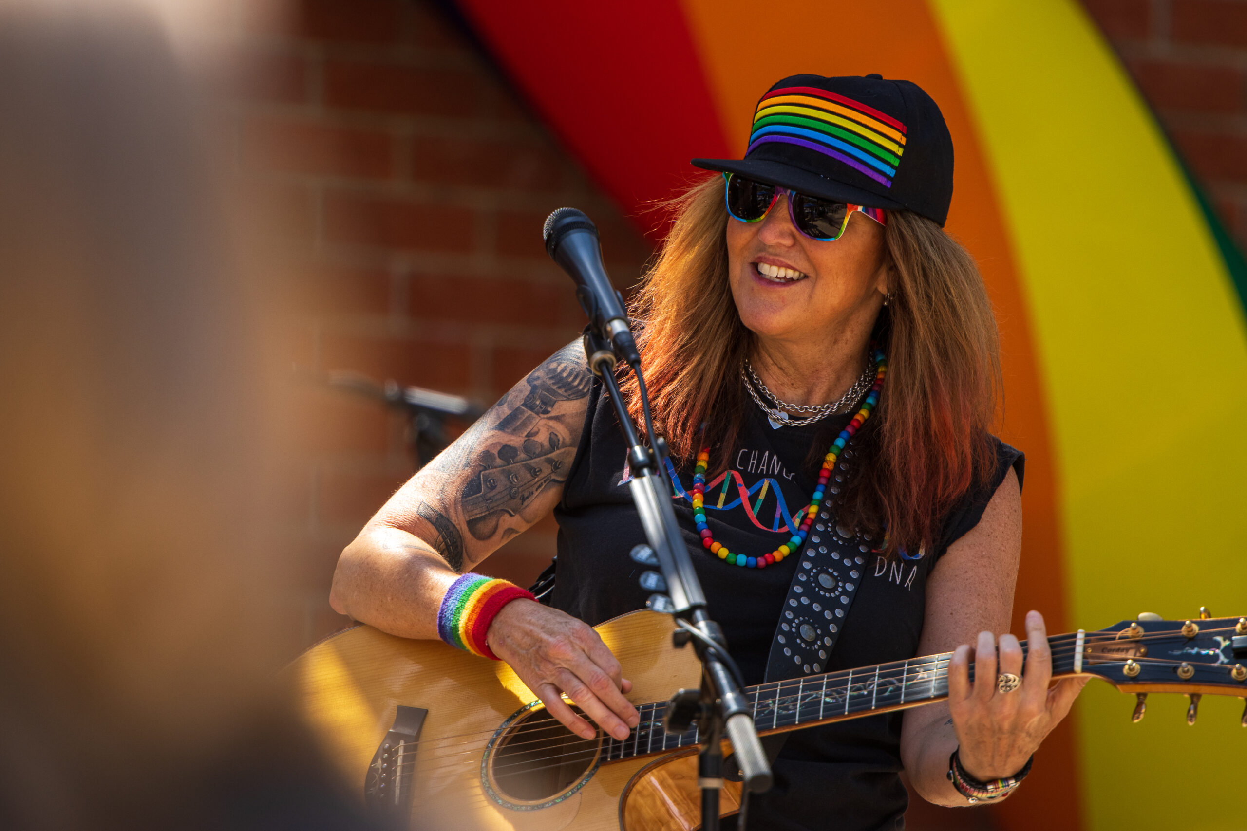 Woman wearing black trucker cap with rainbow and sleeveless black T-shirt plays acoustic guitar on stage.