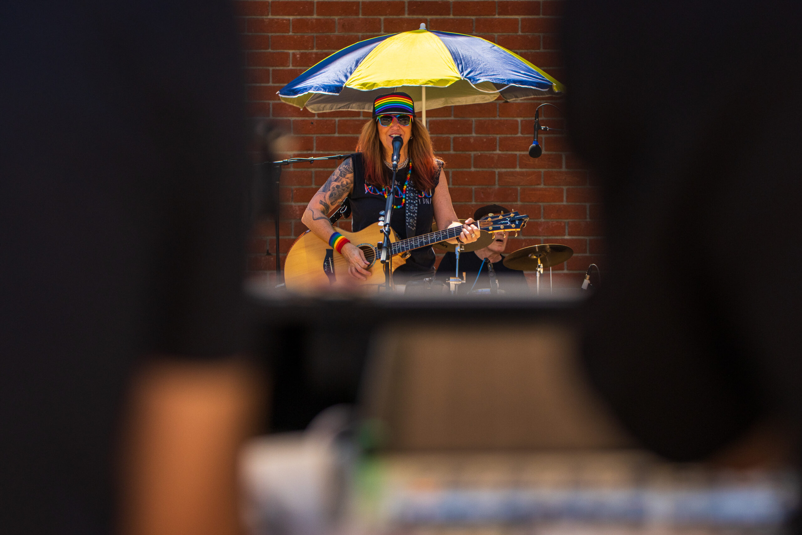 Long shot of woman wearing black trucker cap with rainbow and sleeveless black T-shirt plays acoustic guitar on stage taken from between two sound engineers and the mixing board.
