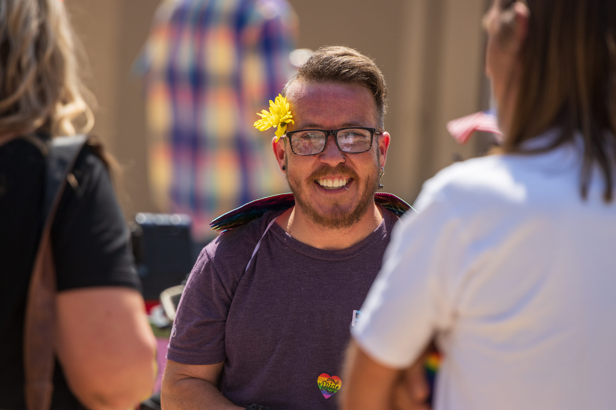 Trans-male wearing dark purple T-shirt, iridescent rainbow unicorn wings, and dark rimmed glasses with a yellow daisy perched behind the right ear smiles generously at the camera while talking to two friends.
