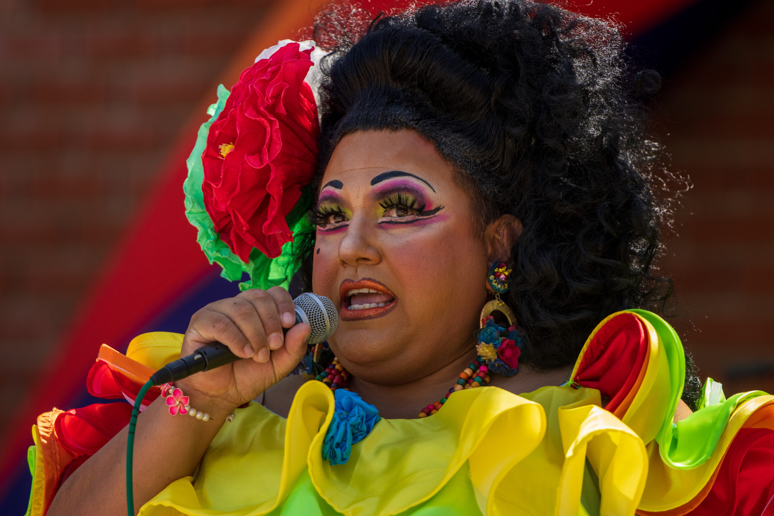 Dressed in a rainbow tiered dress with carnations in their hair, drag queen Kay Sedia addresses the attendees at Pride Fest Fullerton 2023 from the main stage.