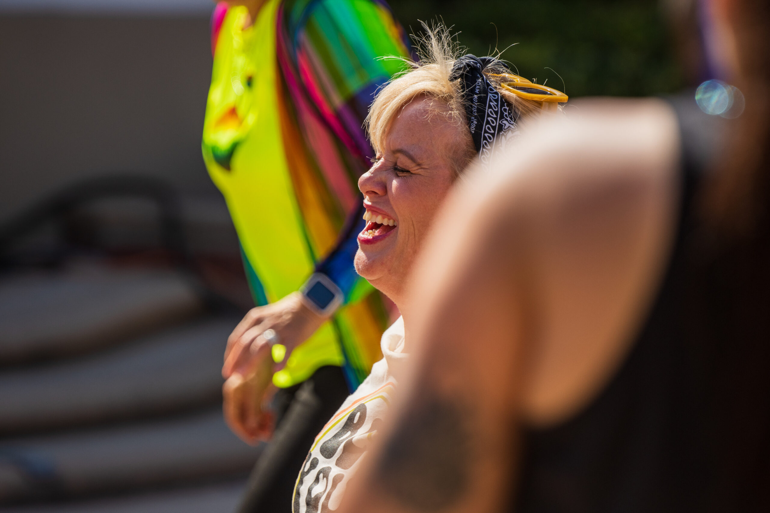 Woman with blonde hair wearing a black bandana and yellow oversized sunglasses on head laughs while being partially obscured by a shoulder of an out of focus woman with black tank top.